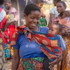Adult carrying a child outdoors among community members.