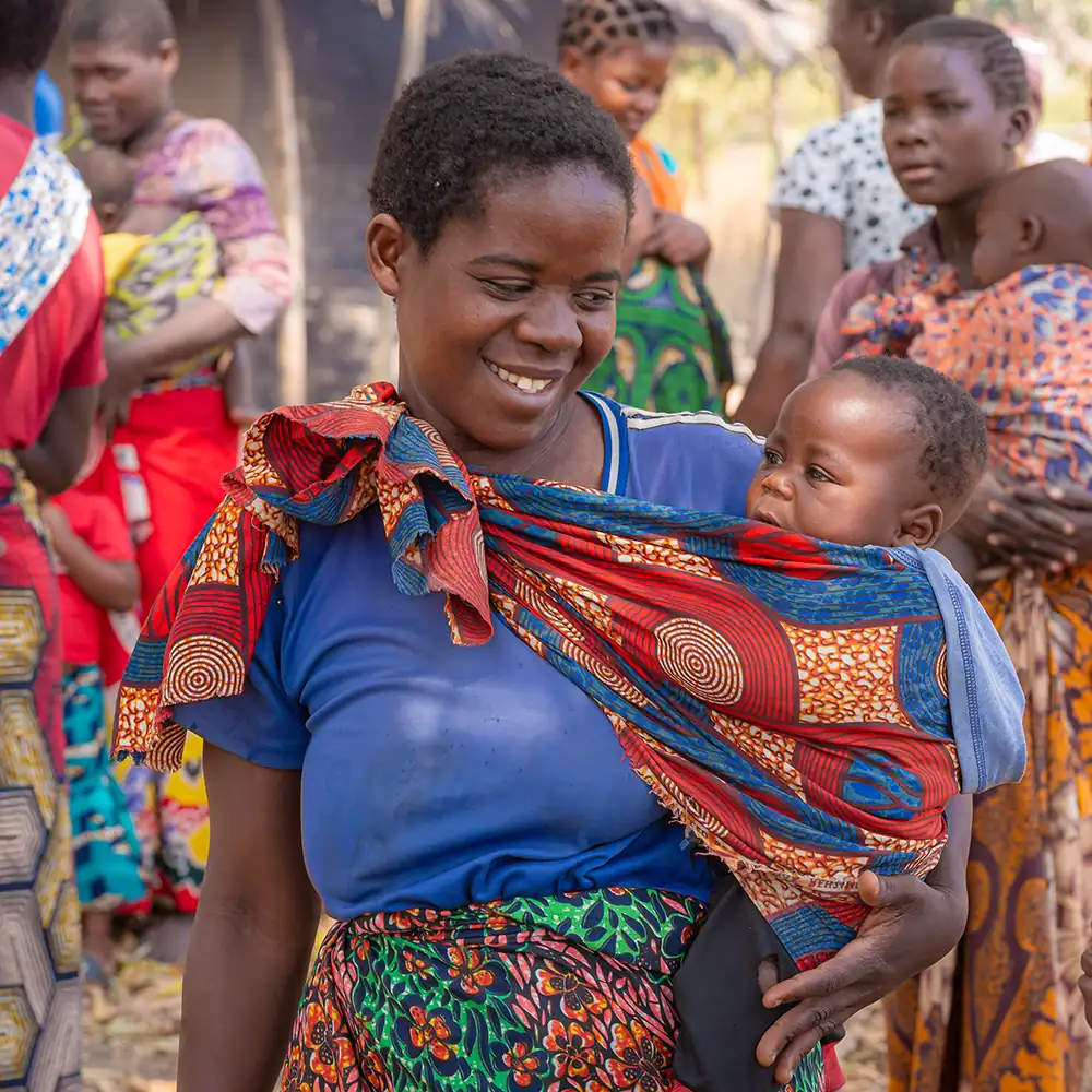 Adult carrying a child outdoors among community members.