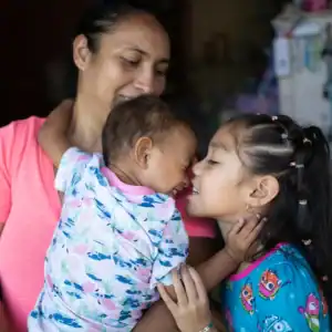 Woman holding a baby while a young child embraces them inside a family home.